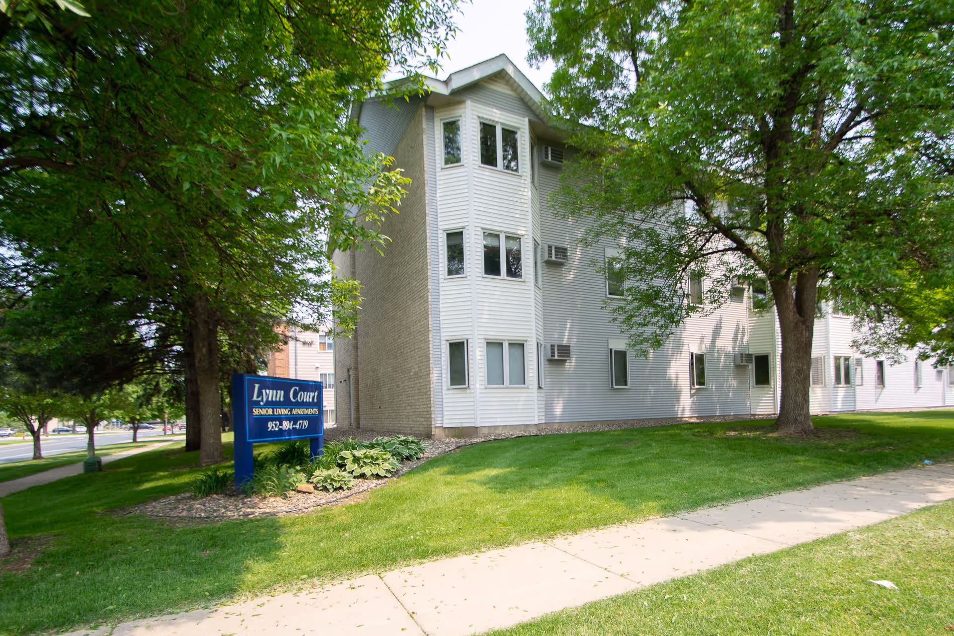 Exterior view of Lynn Court senior living apartments building surrounded by green grass and trees, with a blue sign displaying the facility name and phone number.