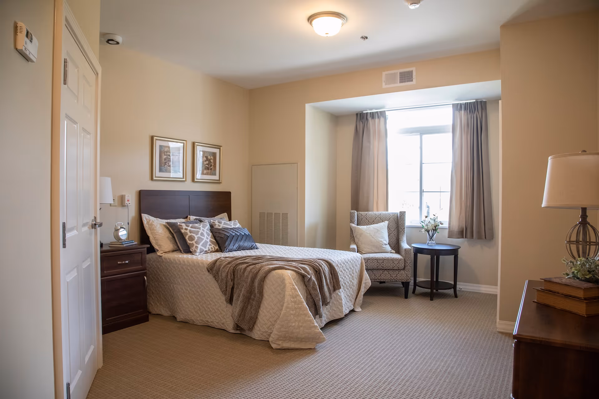 A cozy bedroom with a neatly made bed featuring multiple pillows and a throw blanket. There is a wooden nightstand with a small clock and lamp next to the bed. A patterned armchair with a cushion sits near a window with beige curtains. A wooden dresser with a lamp and decorative books is visible on the right side. The walls are painted light beige and the room is carpeted.