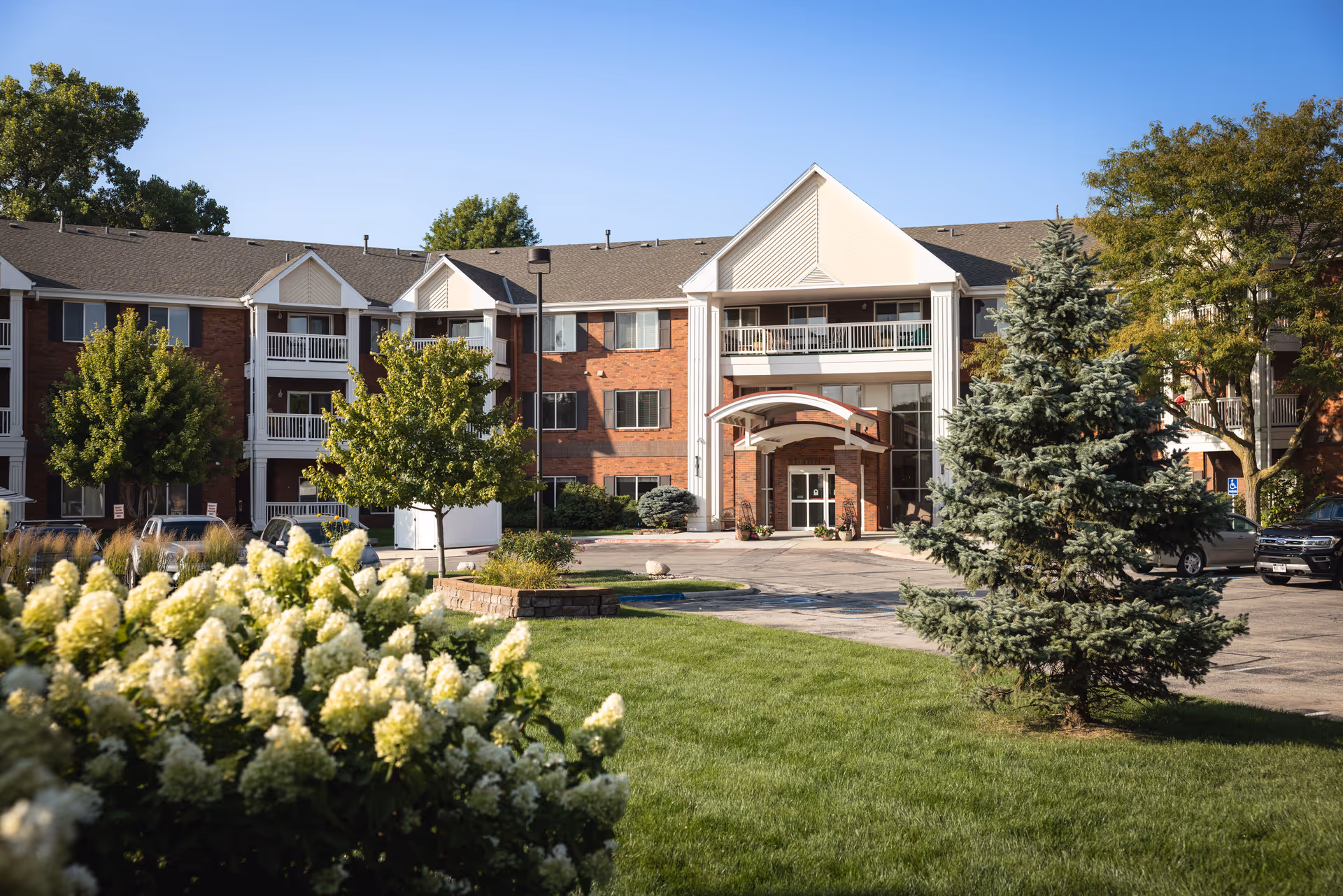 Exterior view of a three-story senior living facility named Bloomfield with a brick facade, white balconies, and a covered entrance. The foreground features green grass, white flowering bushes, and a large evergreen tree under a clear blue sky.