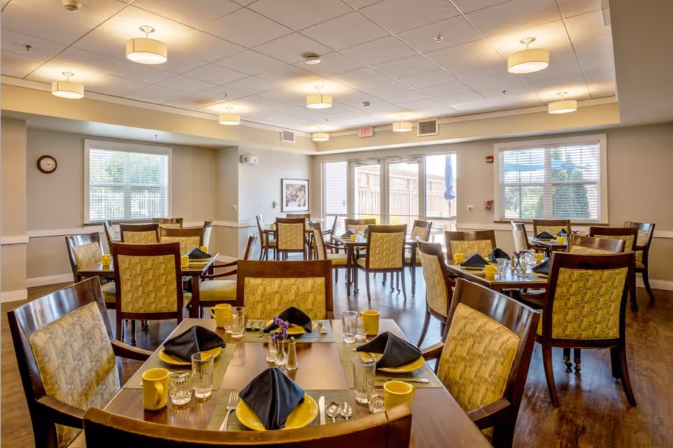 A bright and spacious dining room with multiple wooden tables and chairs arranged neatly. Each table is set with yellow plates, yellow mugs, clear glasses, silverware, and black folded napkins. The room has large windows allowing natural light to enter, beige walls, and ceiling lights providing additional illumination. There is a door leading outside and a framed picture on one wall.