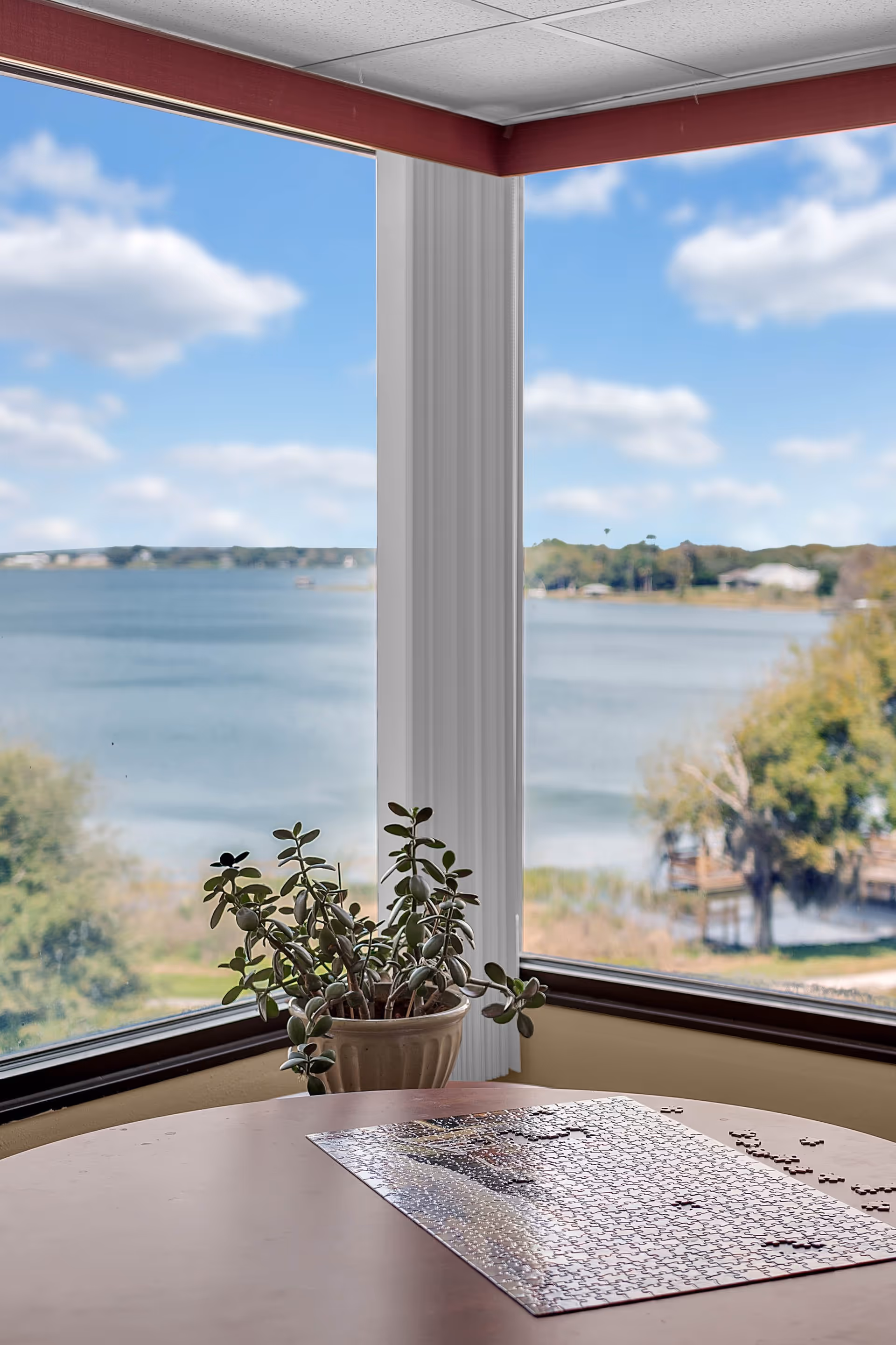 A round table with a partially completed jigsaw puzzle on it, placed near large windows that offer a scenic view of a lake and trees outside. A potted plant sits on the windowsill.