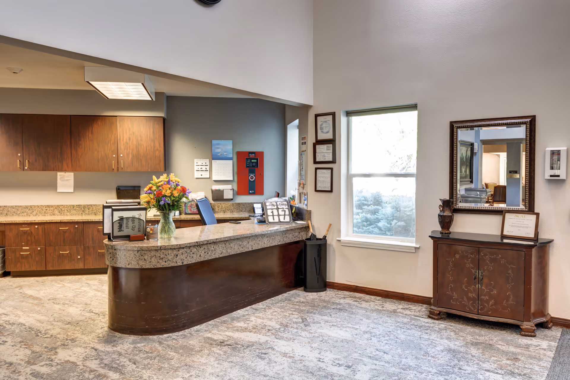 Reception area with a curved granite countertop desk, a vase of colorful flowers, office supplies, and a computer monitor. Behind the desk are wooden cabinets and a wall with a fire alarm and calendar. To the right, there is a window with natural light, a decorative wooden cabinet with a framed mirror above it, and some framed certificates on the wall.