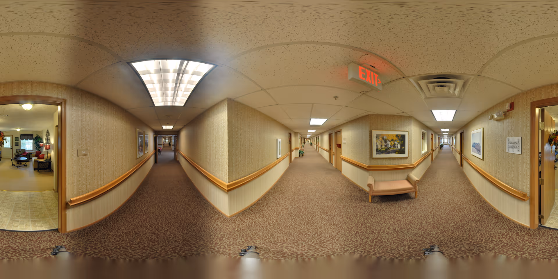 A long, carpeted hallway in a senior living facility with beige walls and wooden handrails on both sides. The ceiling has fluorescent lighting panels and an illuminated exit sign. Several framed paintings hang on the walls, and there is a small bench along one side. Doorways lead to rooms on either side of the hallway, one of which shows a glimpse of a living room area with chairs and a window with curtains.