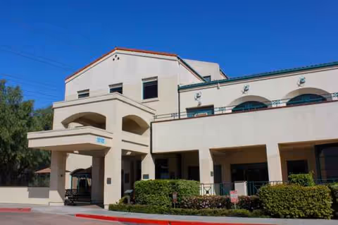 Beige senior living facility exterior with arches, entrance portico, and shrubs.