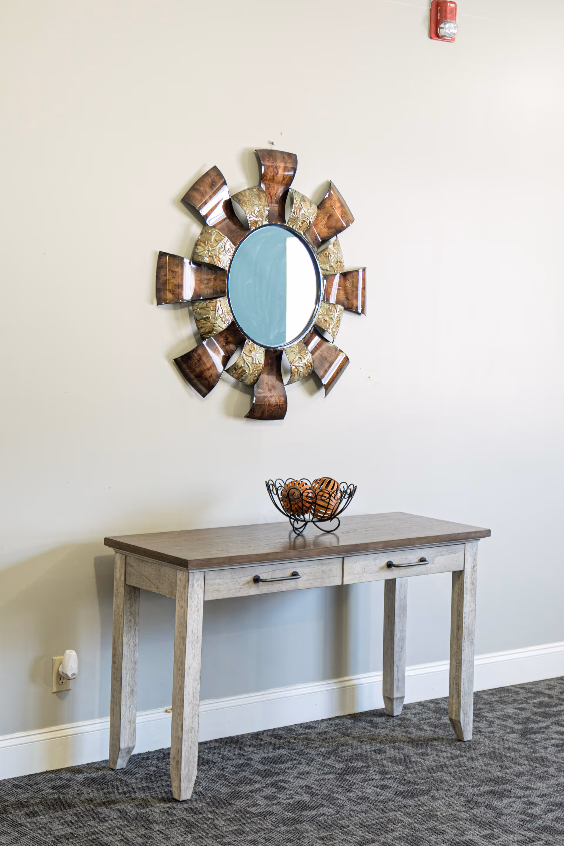 A decorative wooden table with two drawers stands against a light-colored wall. Above the table is a round mirror with a sunburst-style frame made of curved wooden and metallic segments. On the table is a black wire bowl holding decorative wooden balls. The floor is carpeted in a patterned gray design, and a fire alarm is mounted high on the wall.