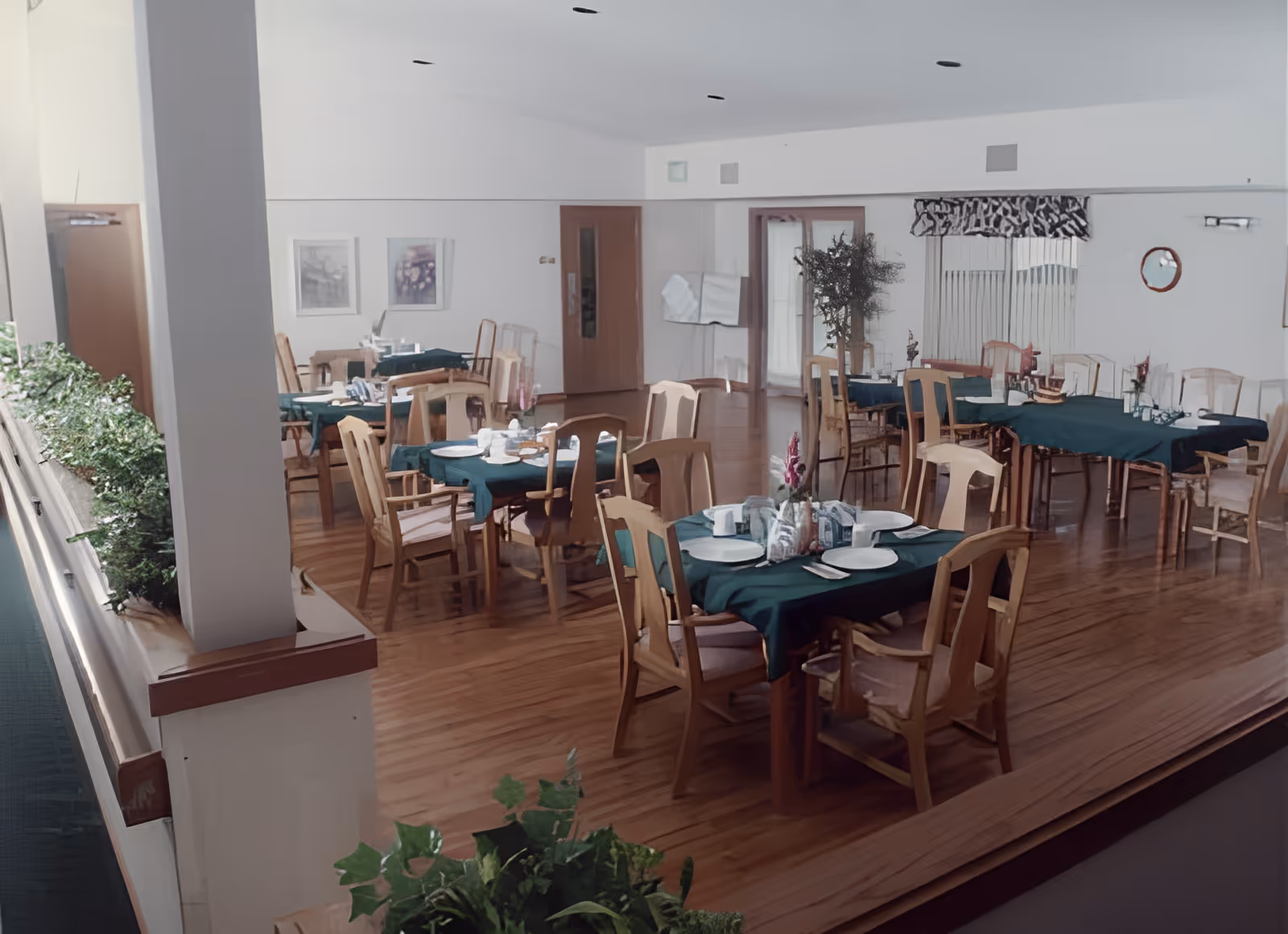 A dining room in an assisted living facility with several wooden tables covered with dark green tablecloths. Each table is set with white plates, cups, and silverware. Wooden chairs surround the tables, and there are plants along the left side of the room. The room has wooden flooring, white walls, and framed artwork on the walls.