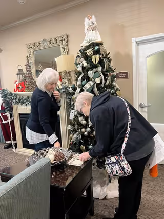 Two elderly women decorating or arranging items near a Christmas tree inside a living room or common area. The tree is decorated with ornaments and topped with an angel. Behind them is a fireplace with holiday decorations and a large ornate mirror above it. A sign on the wall reads 'Business Office.'