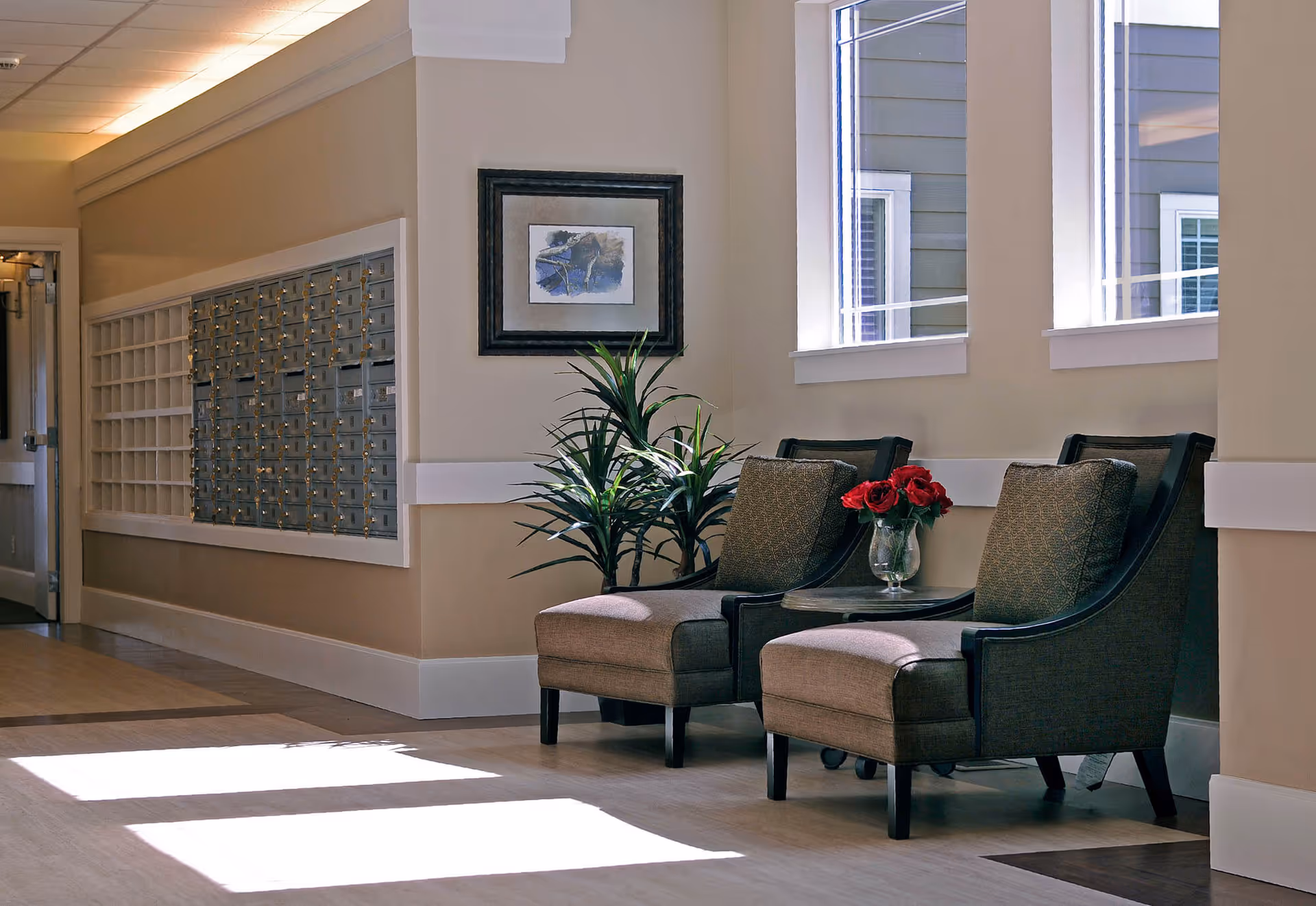 Sunlit senior living lounge with two upholstered chairs, a small table holding a vase of red flowers, a potted plant, and a wall of mailboxes.