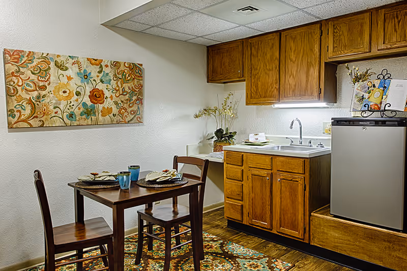 A small dining area and kitchenette in an assisted living facility. The space includes a wooden table set for two with blue cups and napkins, two wooden chairs, wooden cabinets above and below a sink, a small refrigerator, and a colorful floral painting on the wall. The floor has a patterned rug.