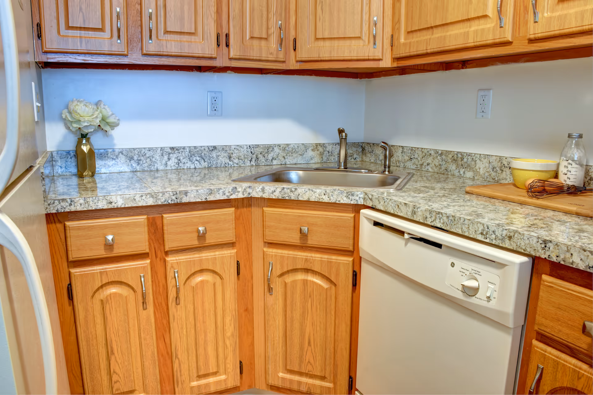 A kitchen corner with wooden cabinets and drawers, a granite countertop, a stainless steel sink with a faucet, a white dishwasher, a small vase with white flowers, a yellow bowl, a glass jar, and a whisk on a wooden cutting board.