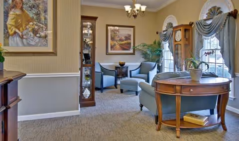 A cozy living room area in a senior living facility featuring light blue upholstered chairs and an ottoman arranged around a small round table. The room has beige walls with white trim, a grandfather clock, framed artwork, and large windows with blue drapes. A wooden table with a potted plant and a book is in the foreground.
