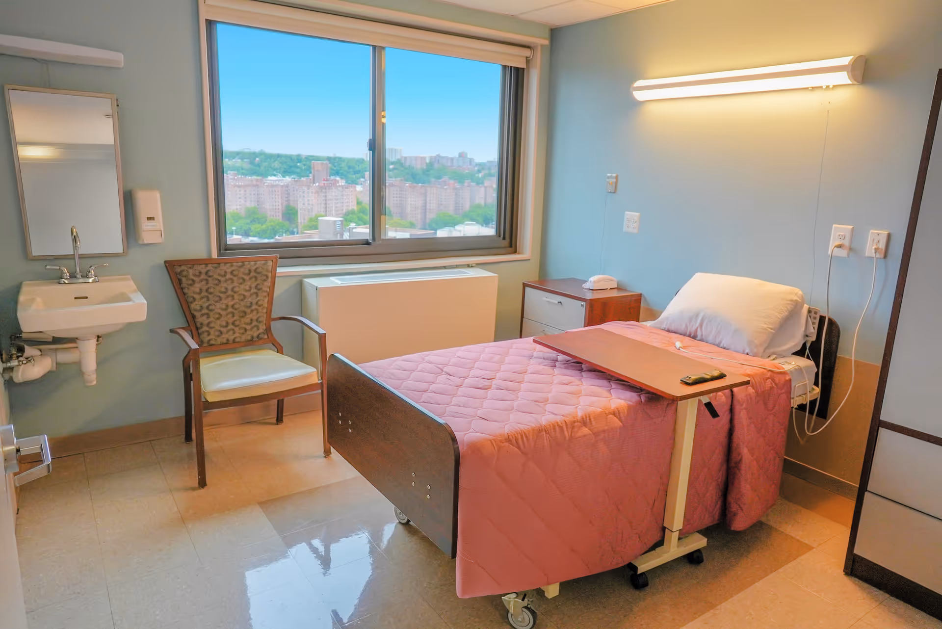 A bright nursing facility patient room with a single bed, overbed table, chair, sink, and a window overlooking the city.