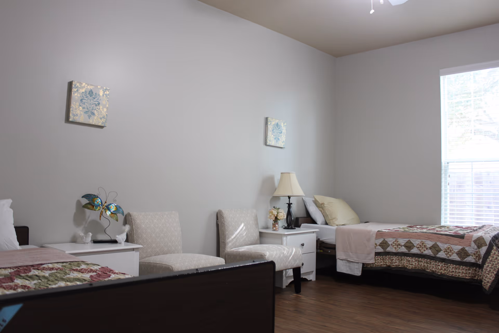 A simple, clean bedroom with two single beds, each with a quilted bedspread. Between the beds are two white nightstands, one with a decorative butterfly sculpture and the other with a lamp and a small flower arrangement. Two upholstered chairs are placed against the wall. The room has light-colored walls, a window with blinds, and wooden flooring.