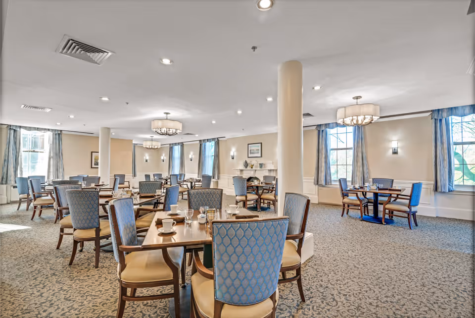 A spacious dining room with multiple wooden tables and cushioned chairs arranged neatly. The room features large windows with blue curtains, soft lighting from ceiling fixtures, and a carpeted floor with a patterned design. The walls are light-colored with decorative molding and framed artwork.