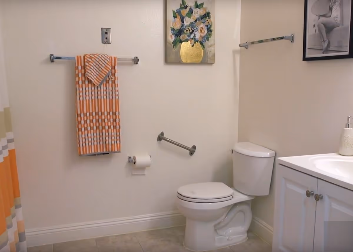 A clean bathroom featuring a white toilet, a white vanity with a sink and soap dispenser, an orange and white patterned towel hanging on a towel rack, a toilet paper holder with a roll of toilet paper, a grab bar next to the toilet, a floral painting above the towel rack, and a black and white framed photo on the wall.