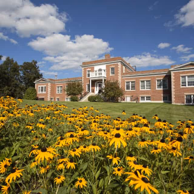 A large brick building with white trim and a small balcony above the entrance, set behind a lawn filled with blooming yellow flowers under a partly cloudy blue sky.