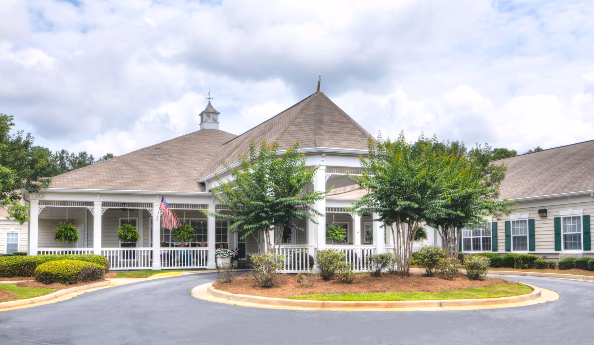 Exterior view of a single-story senior living facility building with a large covered entrance, white railings, hanging plants, and an American flag. The building is surrounded by neatly trimmed bushes and trees under a cloudy sky.