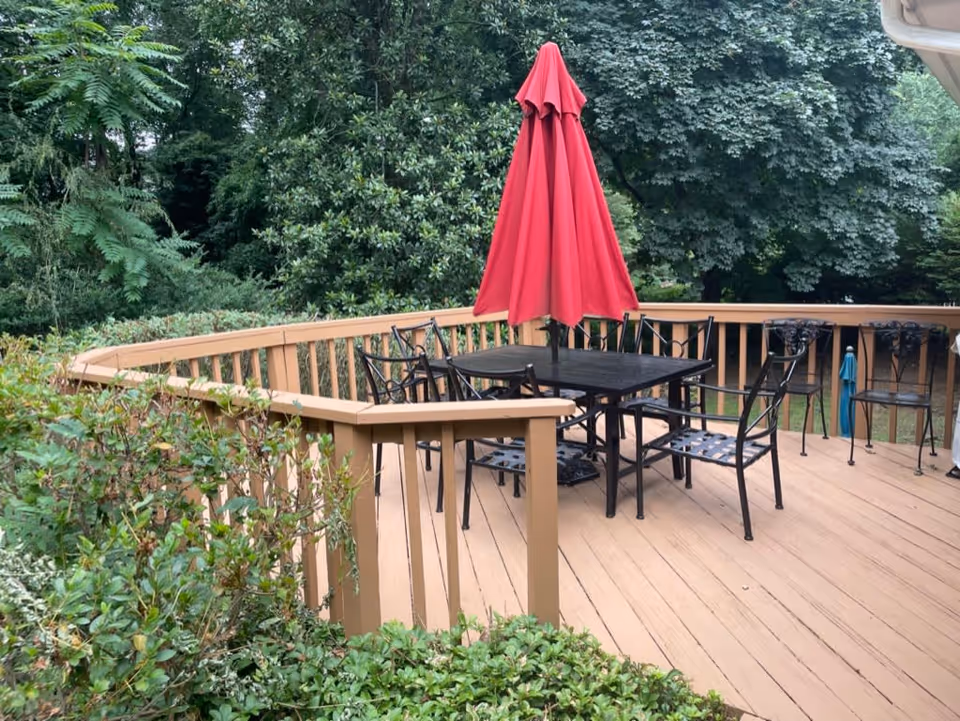 Outdoor wooden deck area with a black metal table and six matching chairs. A closed red patio umbrella is positioned in the center of the table. The deck is surrounded by green bushes and large leafy trees in the background.
