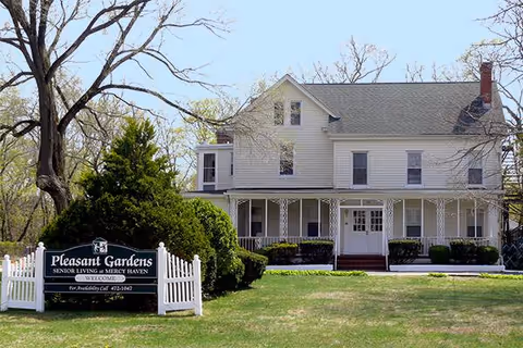 Two-story white house with front porch, green lawn, and Pleasant Gardens Senior Living sign.