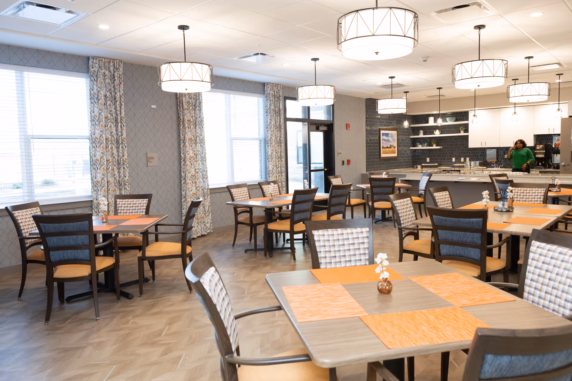 A bright dining room with multiple tables and chairs arranged neatly. Each table has orange placemats and a small decorative vase with white flowers. Large windows with patterned curtains allow natural light to fill the room. Modern pendant lights hang from the ceiling. In the background, there is a kitchen area with white cabinets and a woman in a green top standing near the counter.