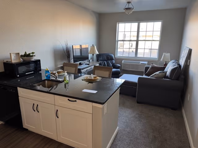 Open-plan living area with a kitchen island in the foreground, gray sofa and armchair by a large window, and a TV on a stand.