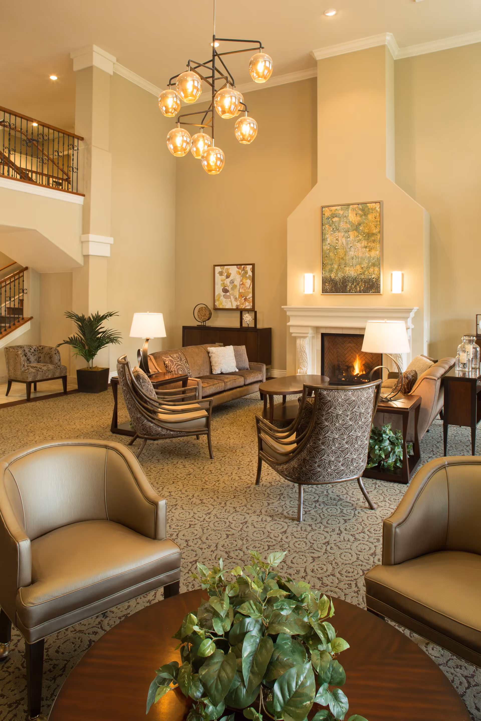 A spacious and well-lit living room area with beige walls and carpeted floor. The room features a fireplace with a painting above it, two sofas, several armchairs, a round wooden coffee table with a green plant on it, and two table lamps. A modern chandelier with multiple bulbs hangs from the ceiling. There is a staircase with a wooden railing visible on the left side.