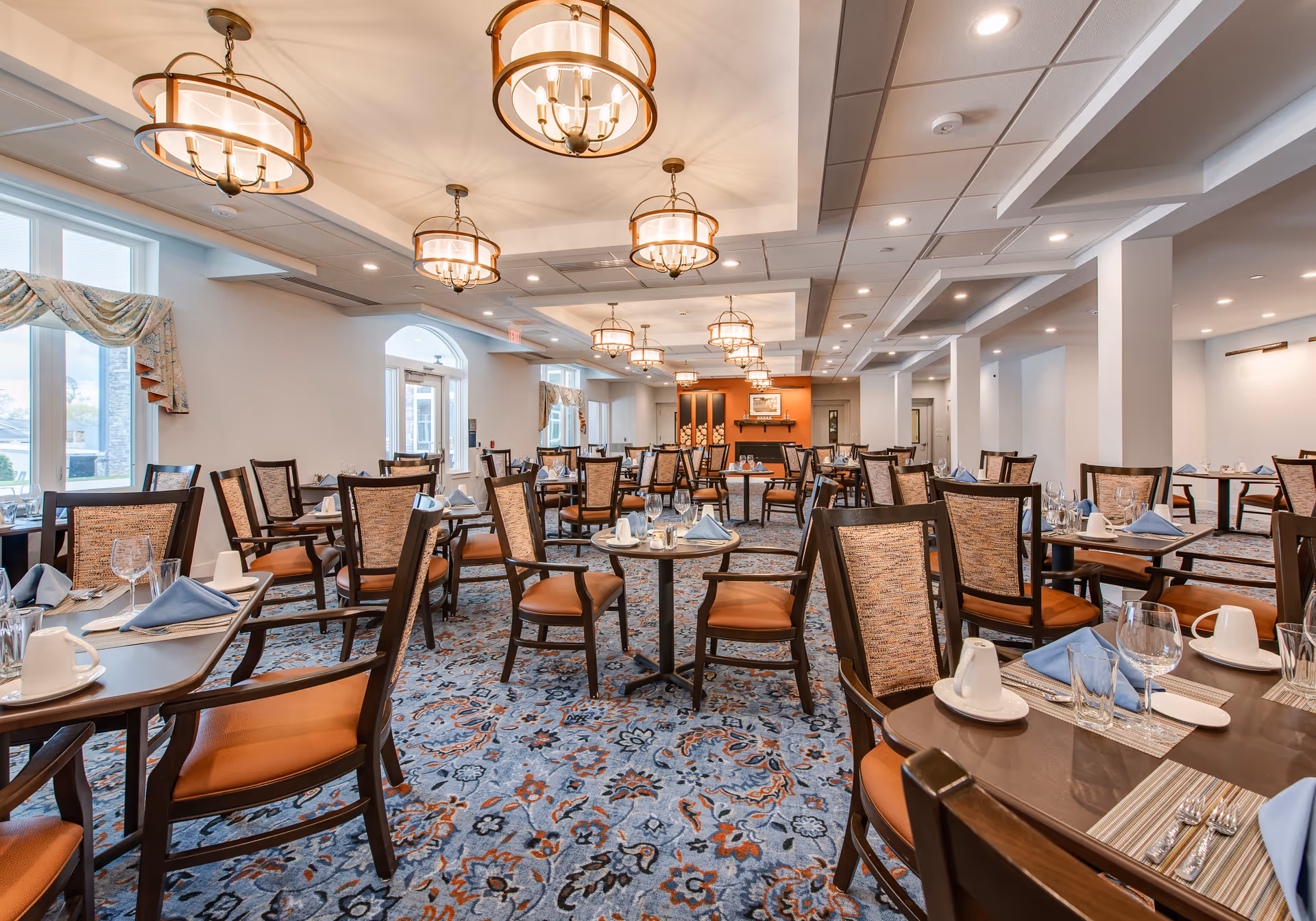 A spacious dining room with multiple tables and chairs arranged neatly. Each table is set with placemats, napkins, glassware, and cups. The room features large windows with decorative curtains, patterned carpet, and several ceiling light fixtures providing warm lighting.