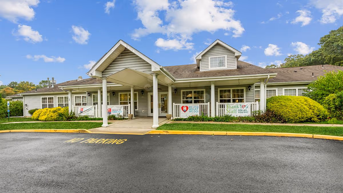 Front exterior view of The Addison of Somers Place facility with a covered entrance, white columns, and a well-maintained lawn with bushes. Signs near the entrance express appreciation for staff and pride in their work. The sky is partly cloudy.