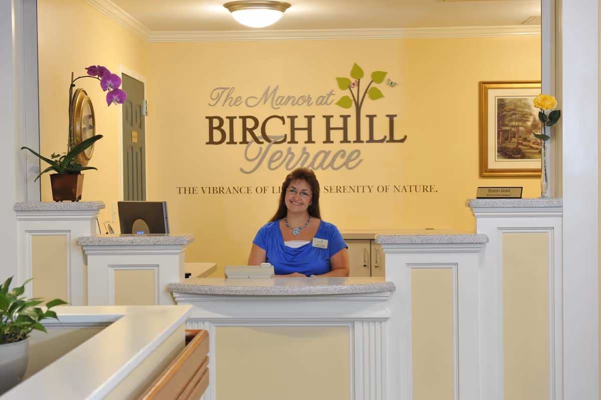 Reception area with a smiling woman standing behind a white counter. Behind her is a wall with the text 'The Manor at Birch Hill Terrace' and the phrase 'The vibrance of life, serenity of nature.' There are potted plants on the counter and a framed picture on the wall.