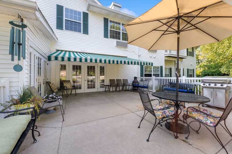 Outdoor patio area at Park Square Manor featuring a round table with floral cushioned chairs under a large beige umbrella, a stainless steel grill, several chairs lined up against the building, a green and white striped awning over glass doors, and potted plants along the edges.