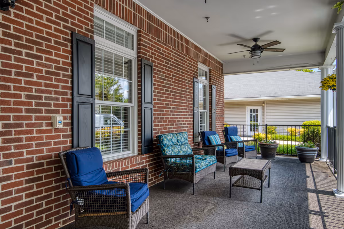 Covered brick-front porch with wicker chairs and cushions, a coffee table, ceiling fan, and potted plants.