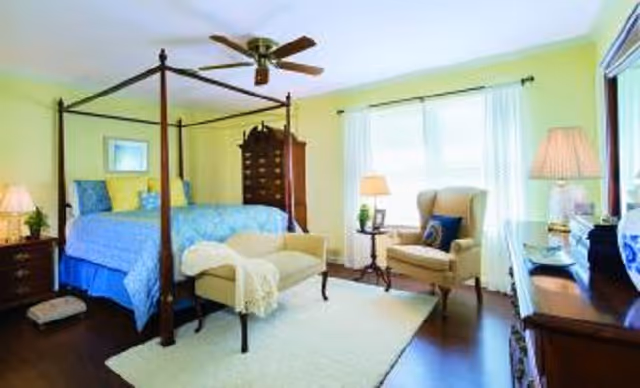 Bright bedroom with a four-poster bed, armchair by a large window, dresser and ceiling fan.