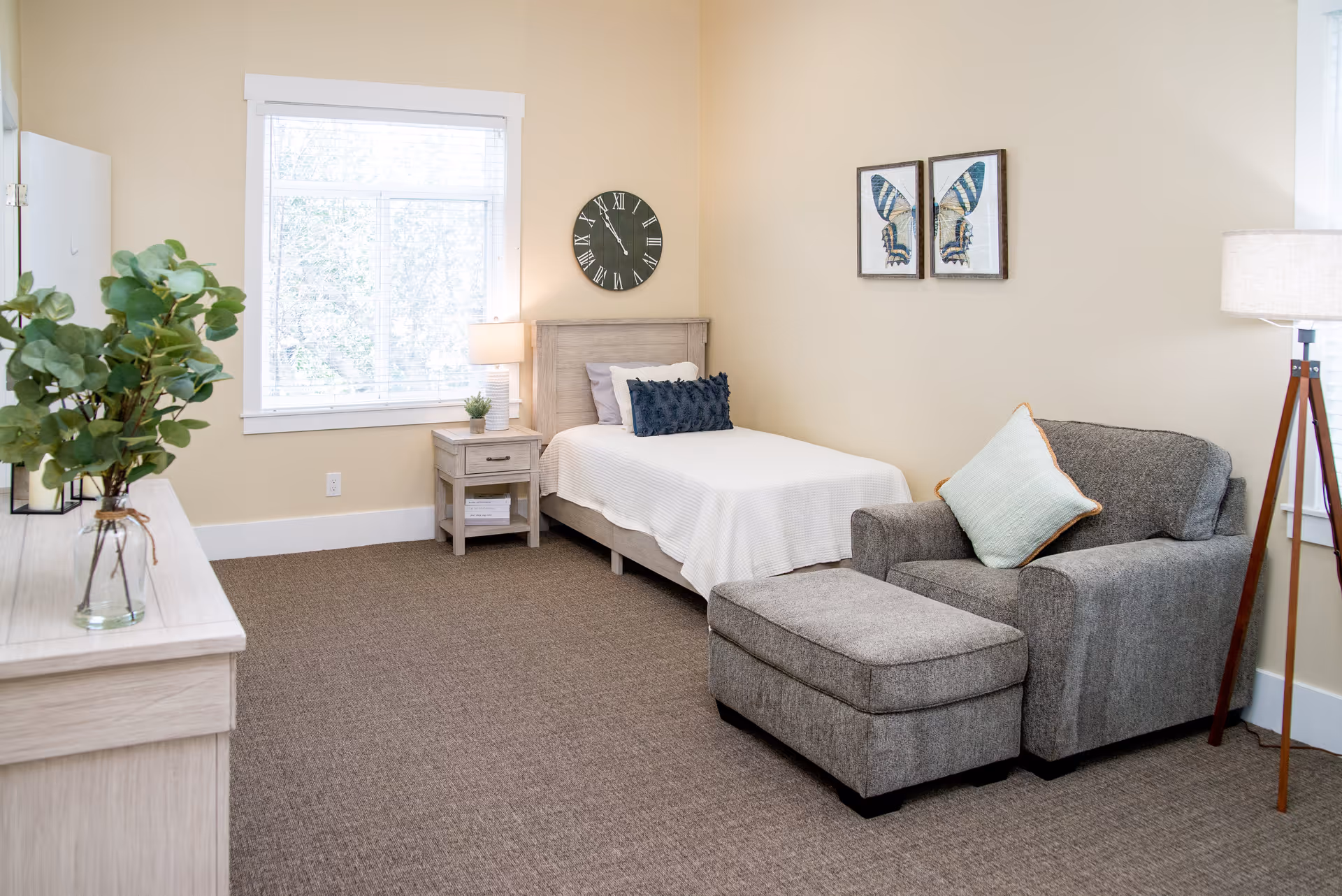 A bright and cozy bedroom in a senior living facility featuring a single bed with white bedding and decorative pillows, a wooden nightstand with a lamp and small plant, a large window with blinds, a gray armchair with a matching ottoman and a light blue pillow, a floor lamp with a tripod base, a wooden dresser with a glass vase containing green foliage, a wall clock above the bed, and two framed butterfly artworks on the wall.