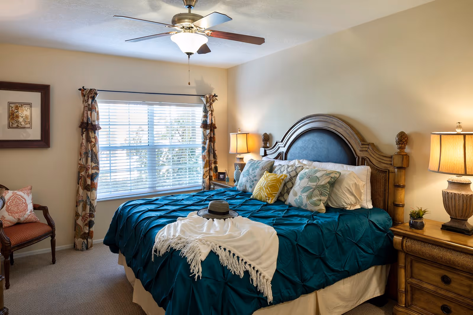 Well-lit bedroom with a large wooden headboard bed dressed in teal bedding and decorative pillows, flanked by nightstands and lamps beneath a window with patterned curtains.