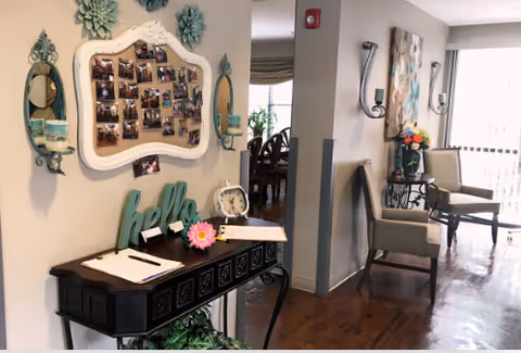 Interior view of a senior living facility hallway with a decorative console table holding clipboards, a clock, and a 'hello' sign. Above the table is a large ornate mirror with photos pinned on it, flanked by two small mirrors and wall sconces. Further down the hallway are chairs, a small table with flowers, and wall decorations, with natural light coming through a window with vertical blinds.