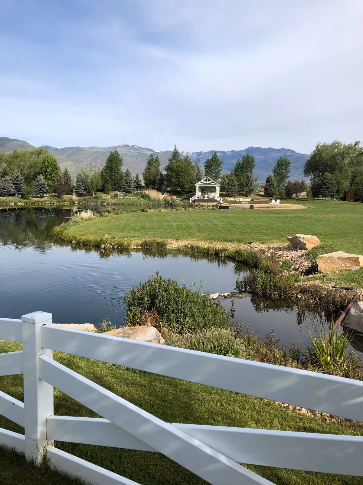A white fence overlooks a pond and grassy lawn with a small white gazebo, trees, and mountains in the background.