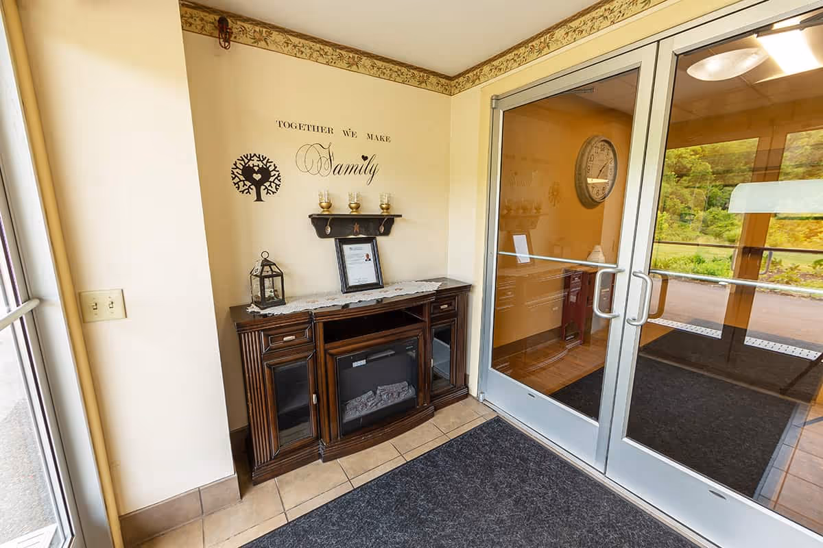 Interior corner of a facility entrance area with glass double doors leading outside. A wooden cabinet with a small electric fireplace is against the wall, decorated with a lantern, framed document, and three candle holders on a small shelf above. The wall has a decal that reads 'Together we make Family' with a tree graphic. A dark floor mat covers the tiled floor near the entrance.