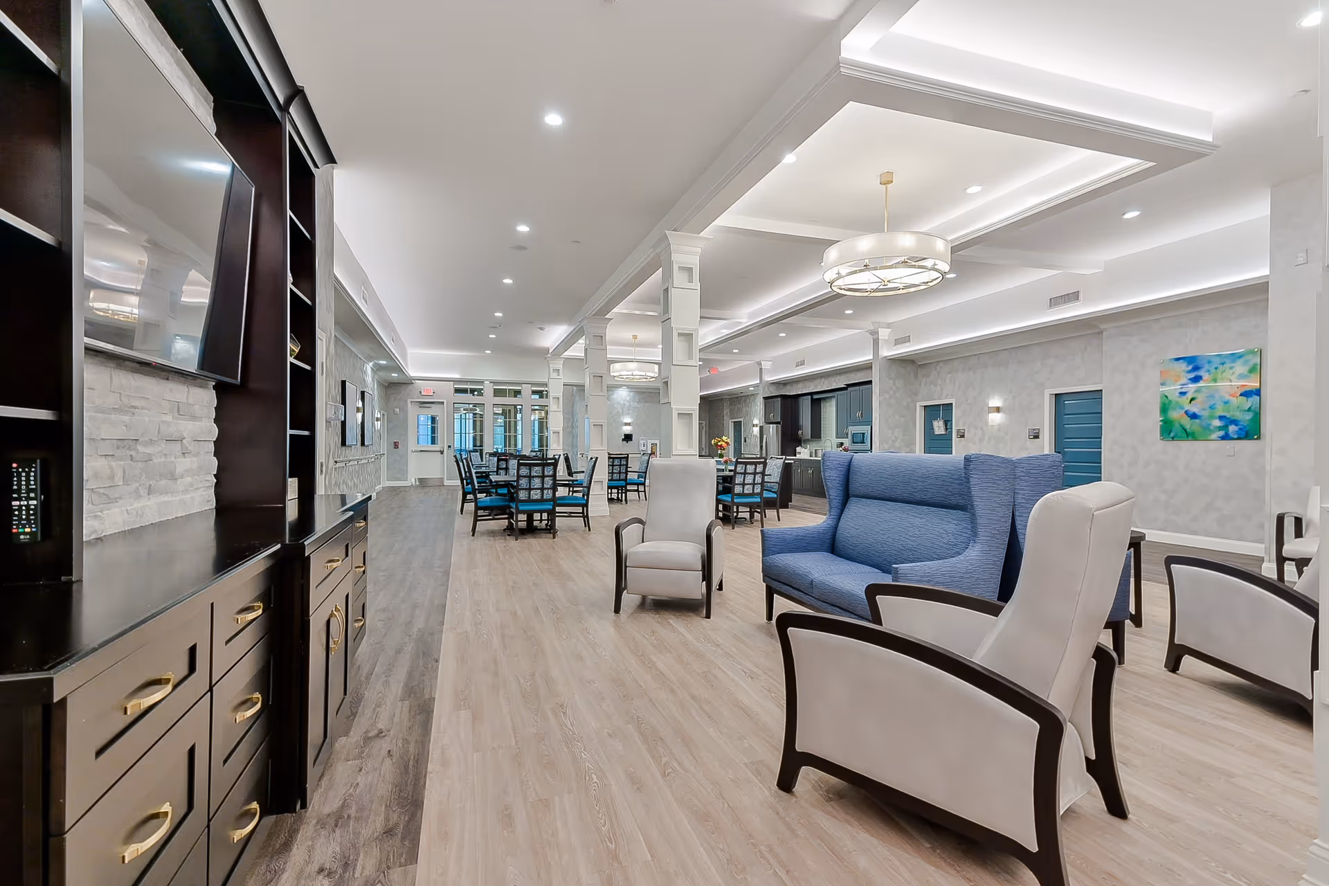 A spacious and modern senior living common area featuring a seating arrangement with blue and beige armchairs and a loveseat, a large flat-screen TV mounted on a dark wood entertainment unit, multiple dining tables with chairs in the background, and a kitchen area with dark cabinetry. The room has light wood flooring, white walls with subtle patterns, recessed lighting, and decorative ceiling fixtures.