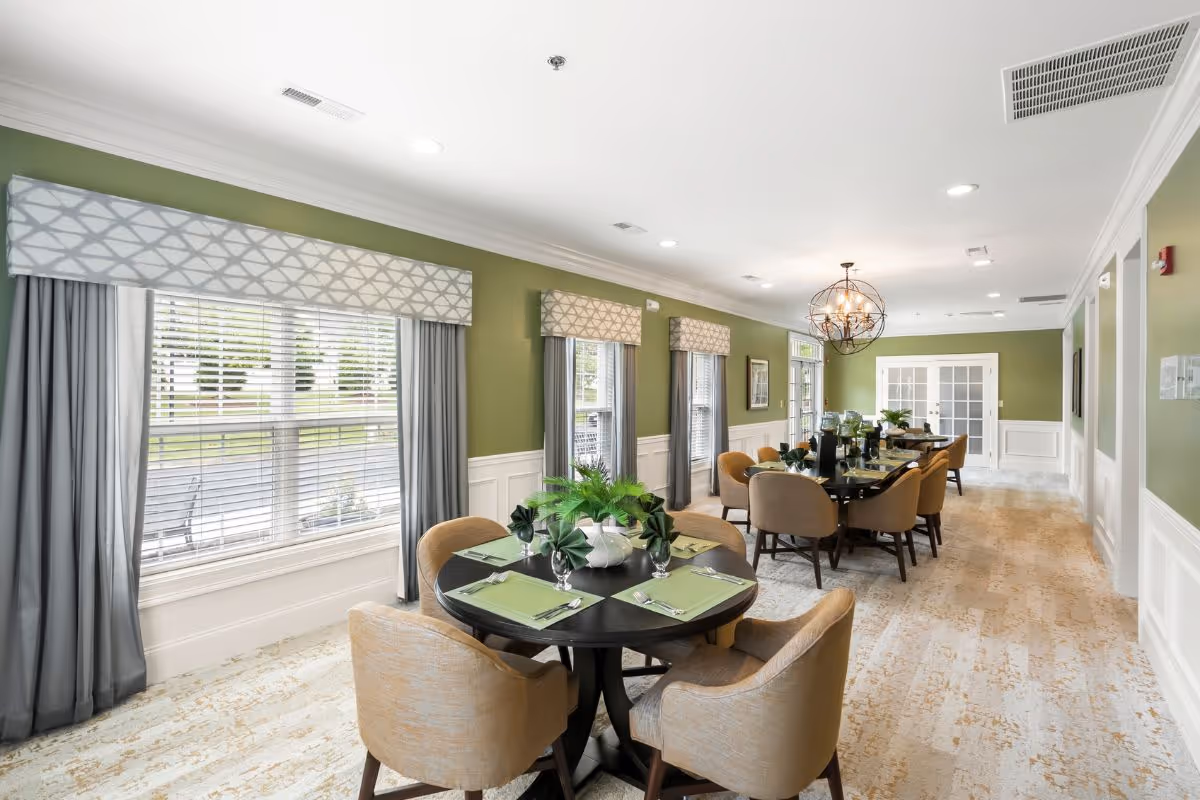A bright dining room with green walls and large windows covered with patterned valances and gray curtains. The room features round and rectangular dining tables set with green placemats, folded napkins, glassware, and decorative plants. Beige upholstered chairs surround the tables, and a modern chandelier hangs from the ceiling. The floor has a light-colored patterned carpet.