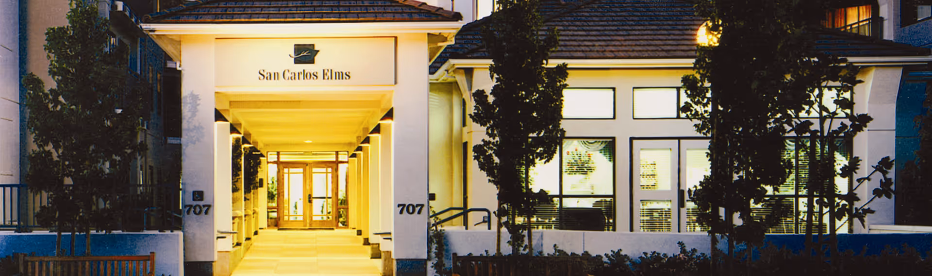 Illuminated entrance of the San Carlos Elms building with a covered walkway leading to glass doors.