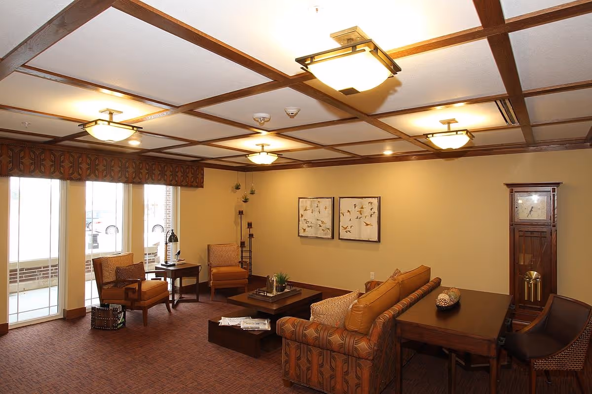 A warm, furnished lounge area with sofas and armchairs centered around a coffee table beneath coffered ceiling lights and large windows.