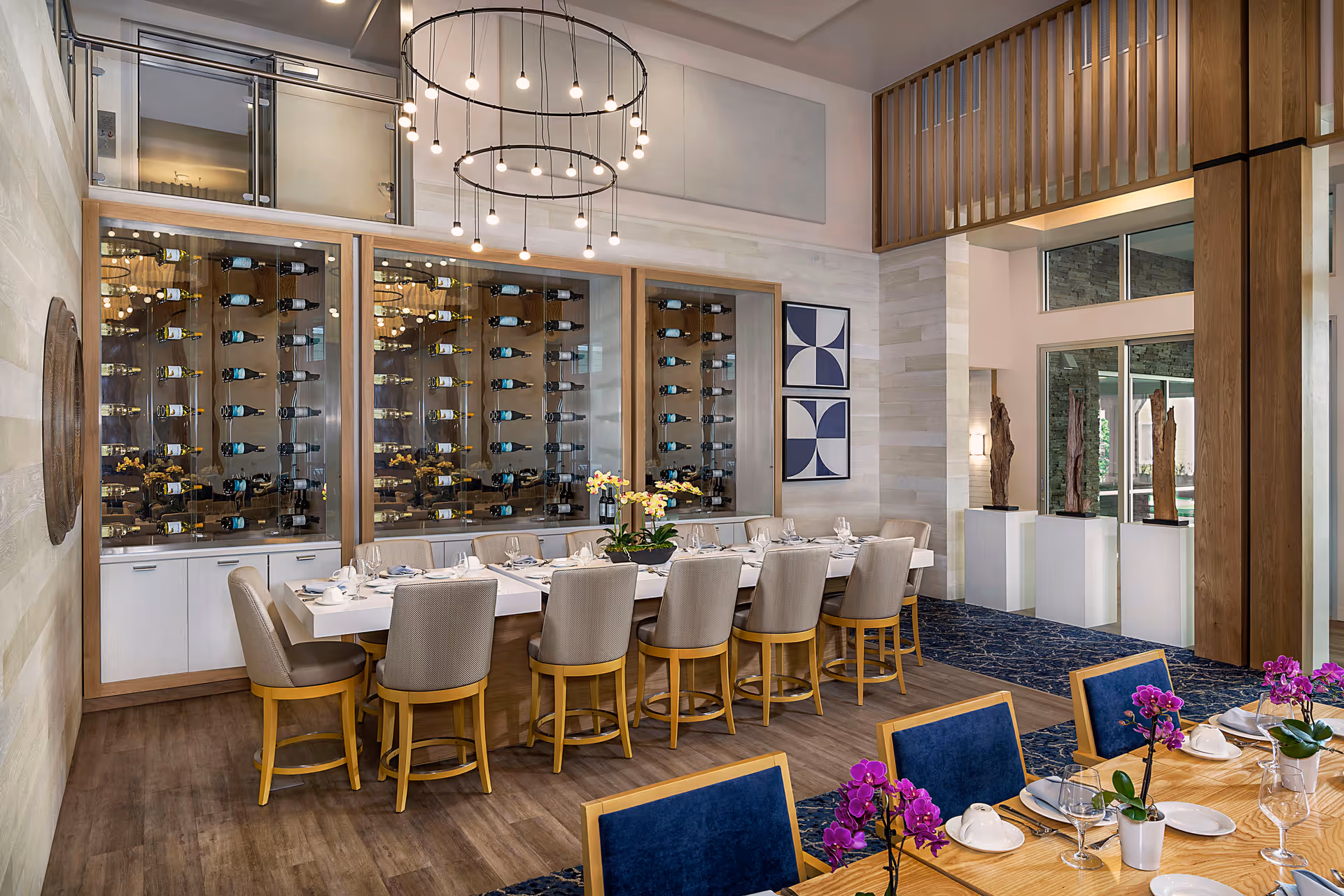 A modern dining room with a long white table surrounded by beige chairs with wooden legs. Behind the table is a glass-enclosed wine rack filled with bottles. The room features contemporary lighting with two circular chandeliers hanging from the ceiling. There are decorative wall art pieces and sculptures on white pedestals near large windows that let in natural light. Another wooden table with blue cushioned chairs and purple orchids in white pots is partially visible in the foreground.