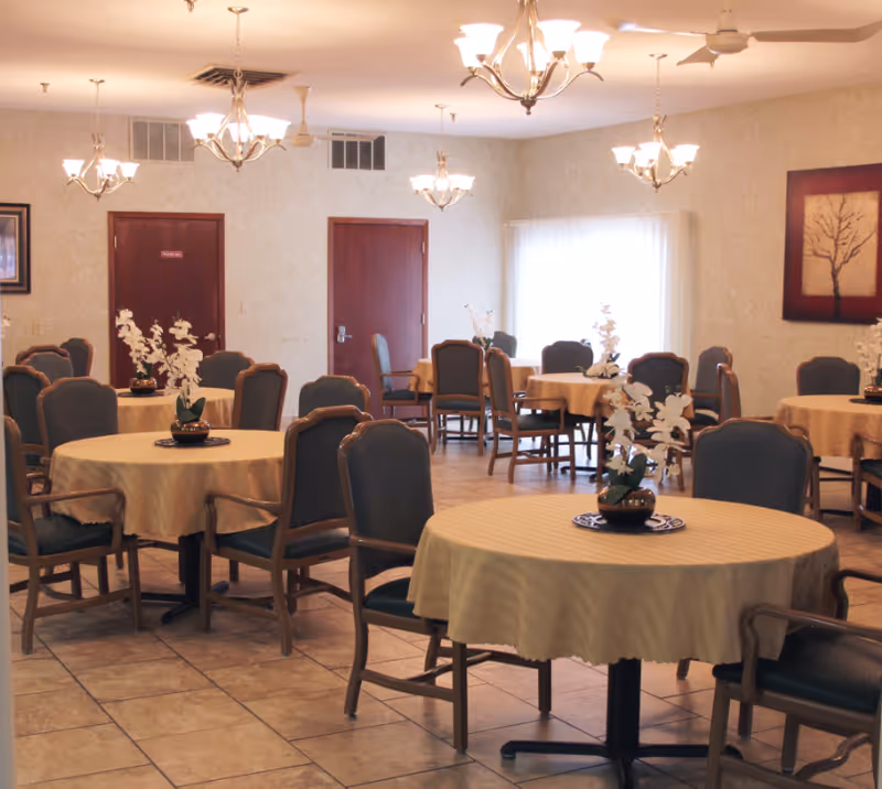 Bright dining room with round tables draped in beige tablecloths, upholstered chairs, chandeliers, and floral centerpieces.