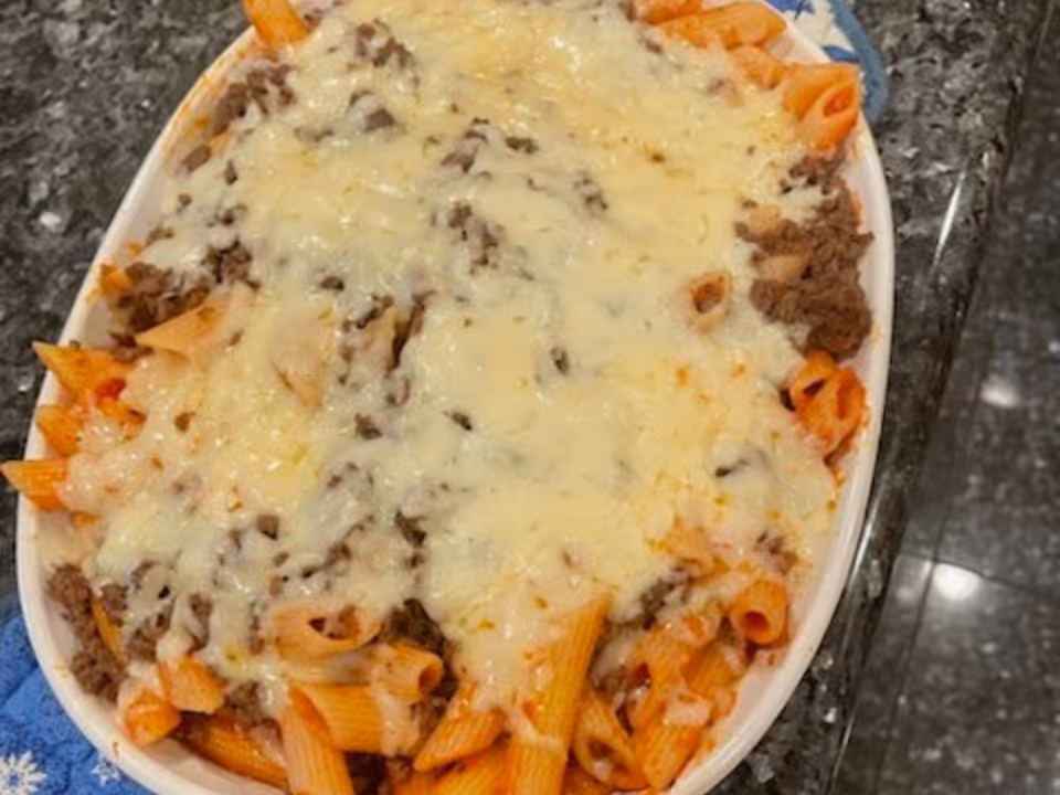 Oval baking dish of baked penne pasta topped with melted cheese and ground meat on a dark countertop.