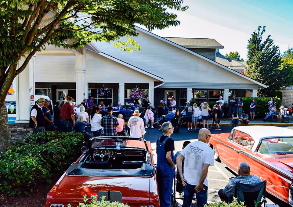 A crowd of people and classic cars gathered outside the front entrance of a senior living facility.