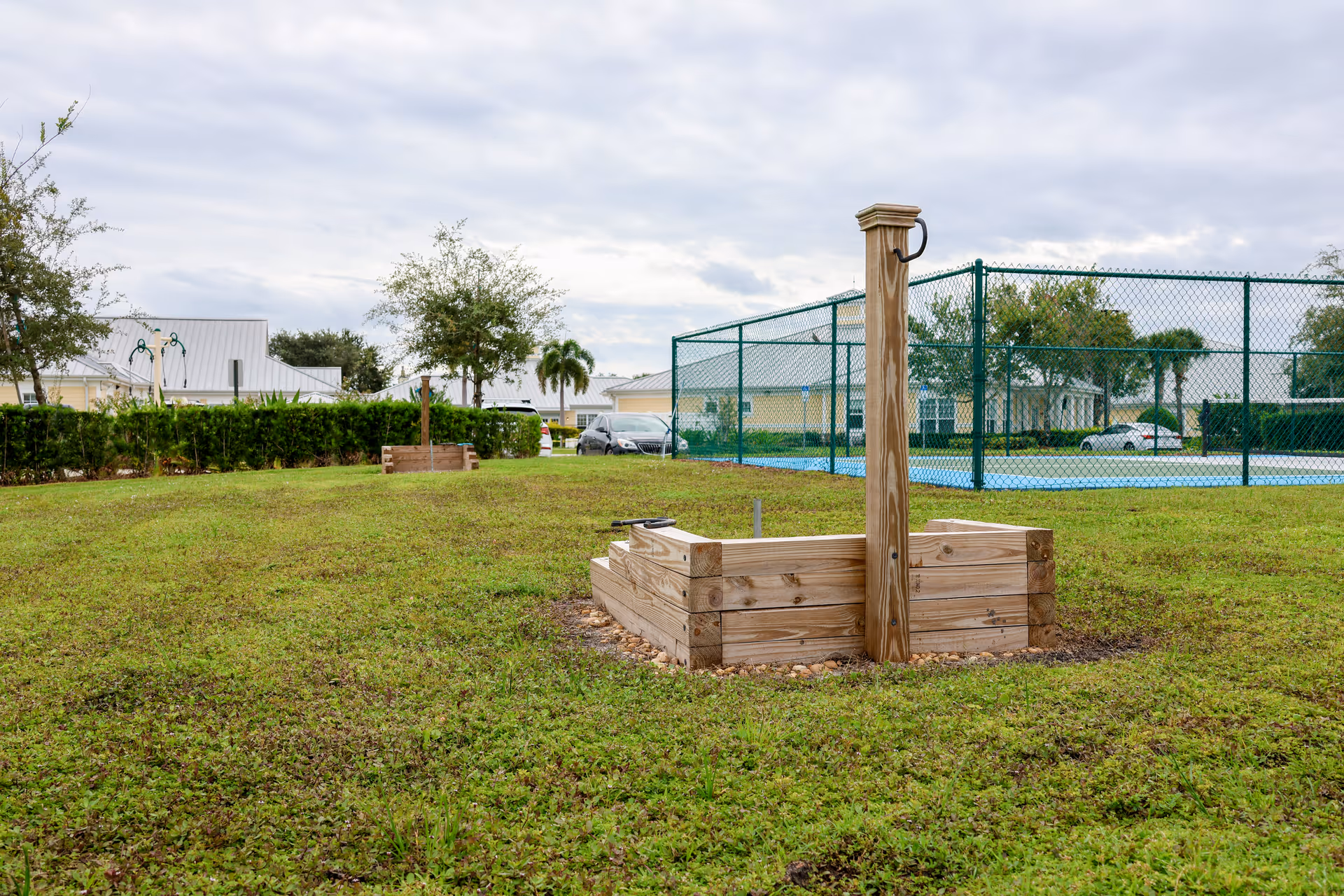 Outdoor area with green grass, two wooden raised garden beds, a tall wooden post with a metal ring, a chain-link fence enclosing a tennis or basketball court, and buildings and trees in the background under a cloudy sky.