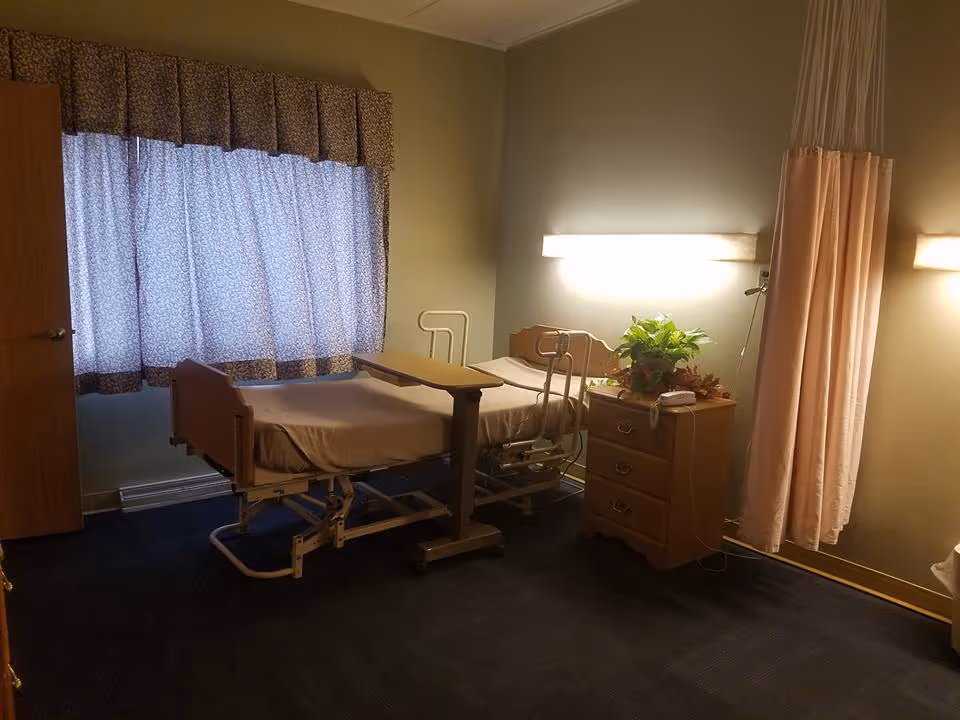 A senior care resident room with an adjustable hospital bed, overbed table, nightstand with a plant, window curtains and a privacy curtain.