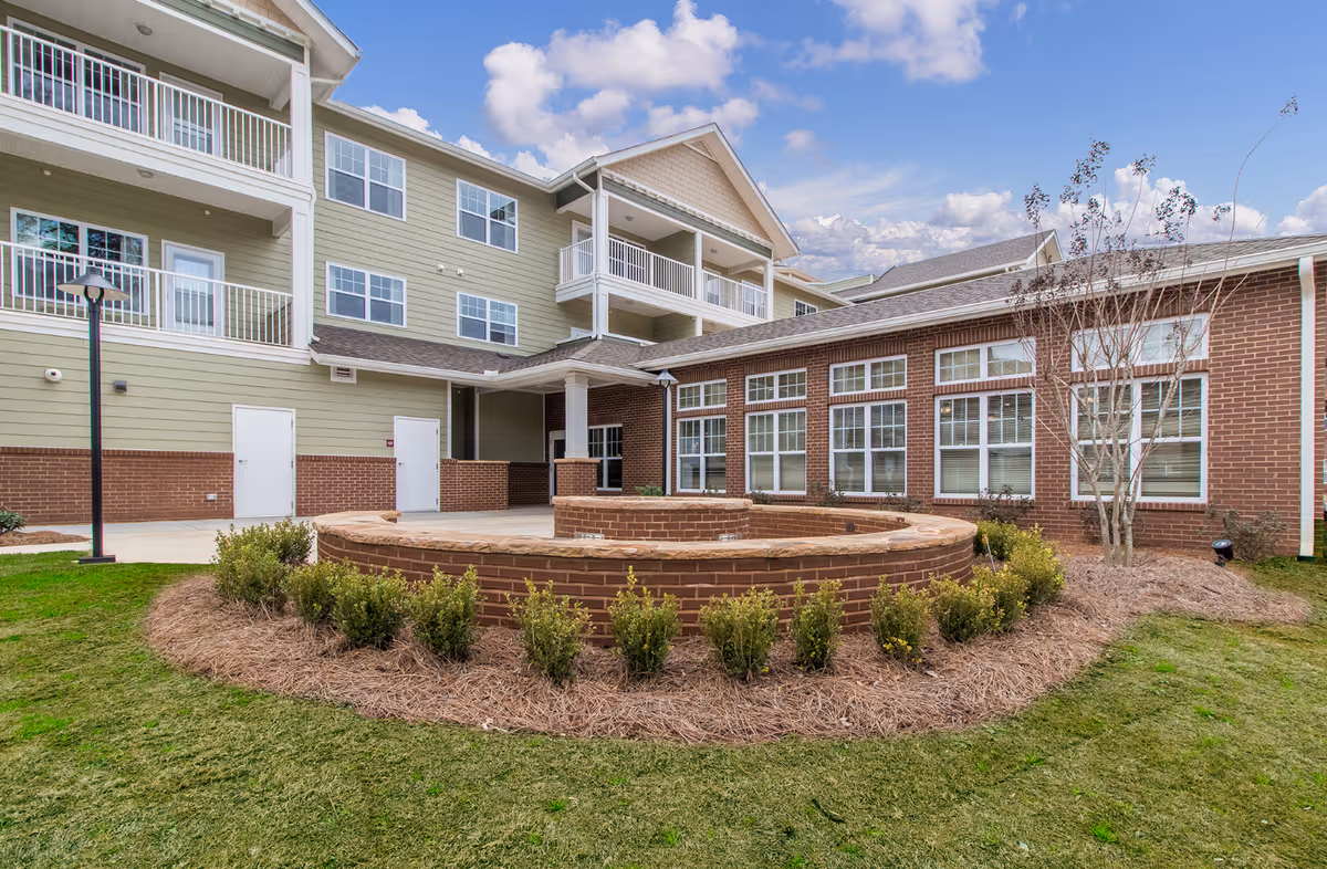 Outdoor view of Linwood Estates Gracious Retirement Living showing a multi-story building with balconies, large windows, and a landscaped garden area with a circular brick planter and small shrubs under a partly cloudy sky.