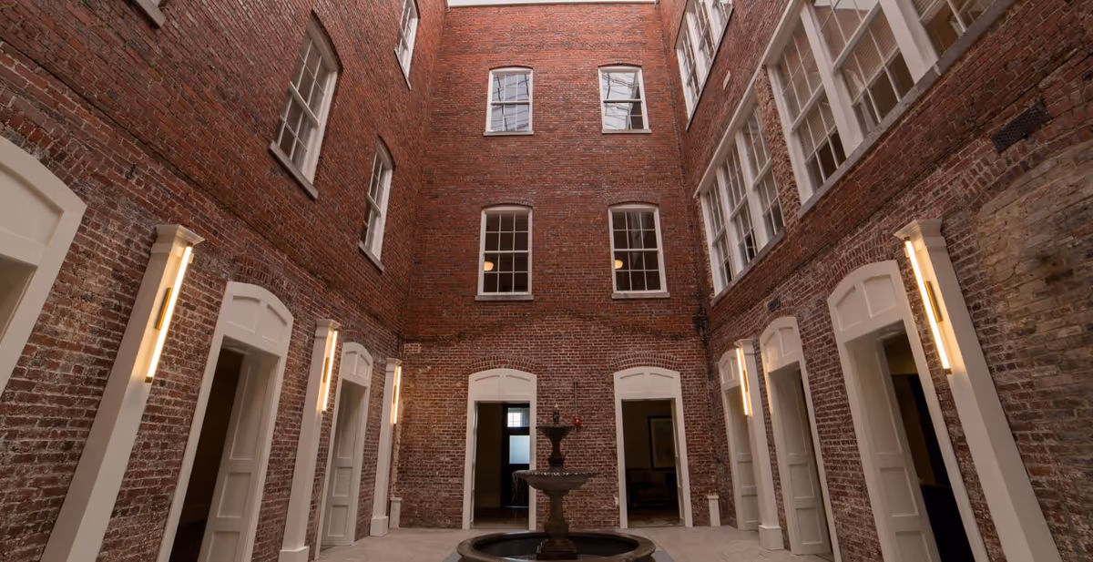 Interior courtyard of Knoxville High Apartments featuring exposed red brick walls, multiple white-framed windows, several doorways with white trim, vertical wall lights, and a central two-tiered water fountain.