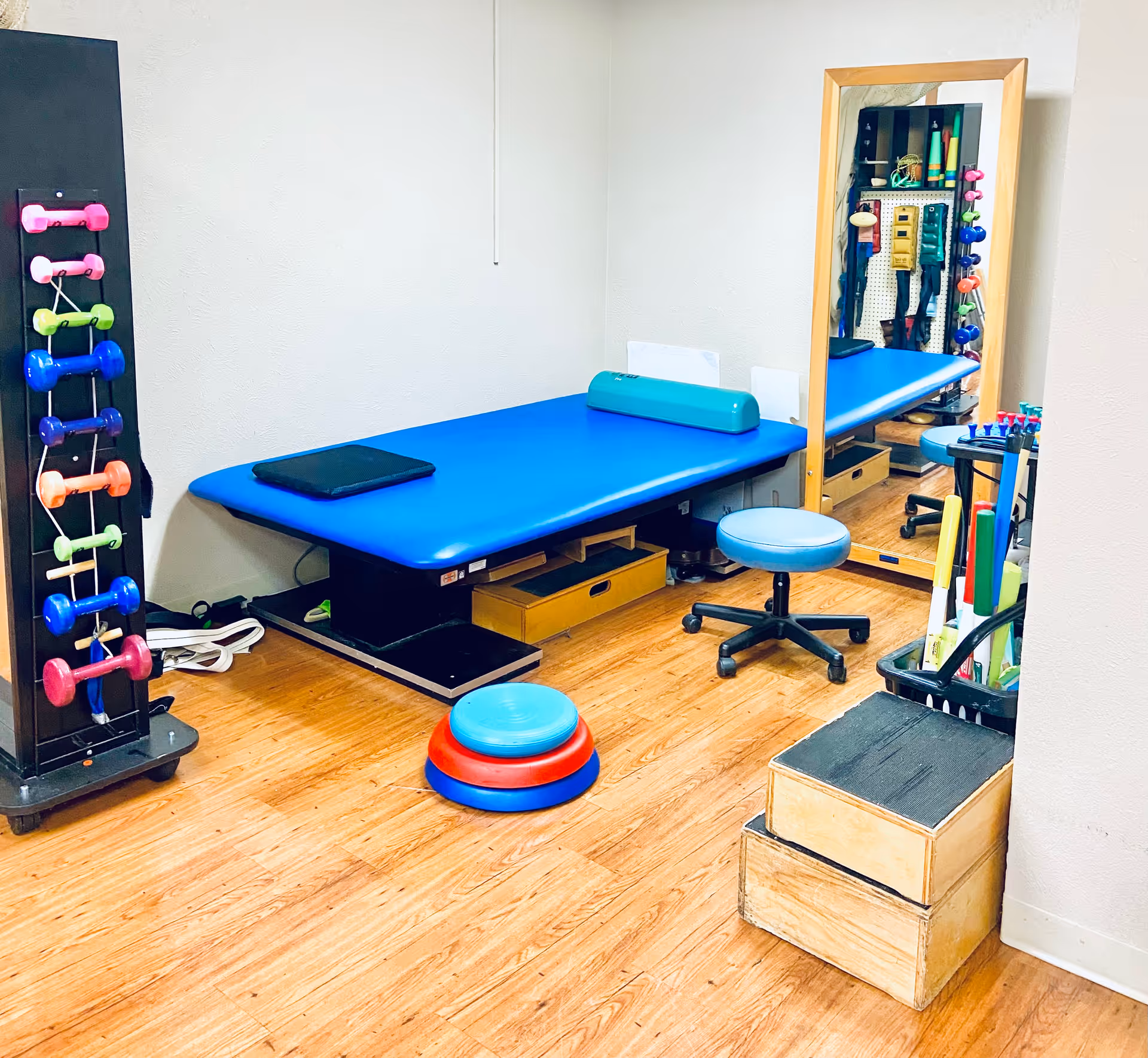 A therapy or exercise room with a blue padded treatment table, a small blue rolling stool, a set of colorful dumbbells on a rack, a large mirror, wooden steps, and various exercise equipment on a wooden floor.